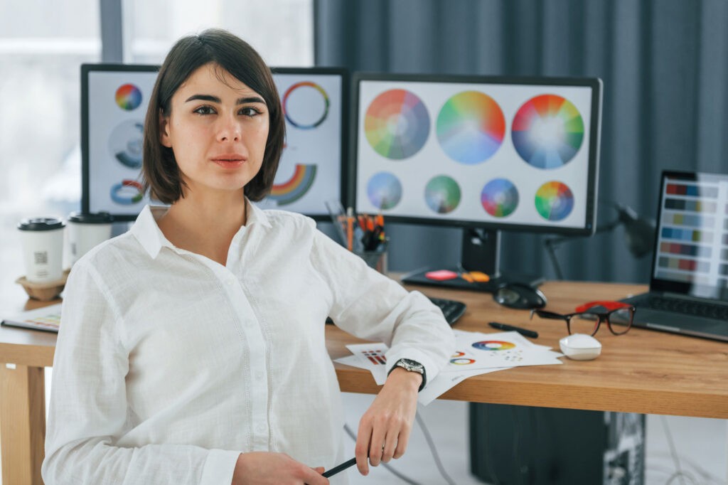 Designer grafica seduta alla scrivania in ufficio, circondata da monitor con ruote cromatiche e schede colore, mentre tiene una penna in mano.