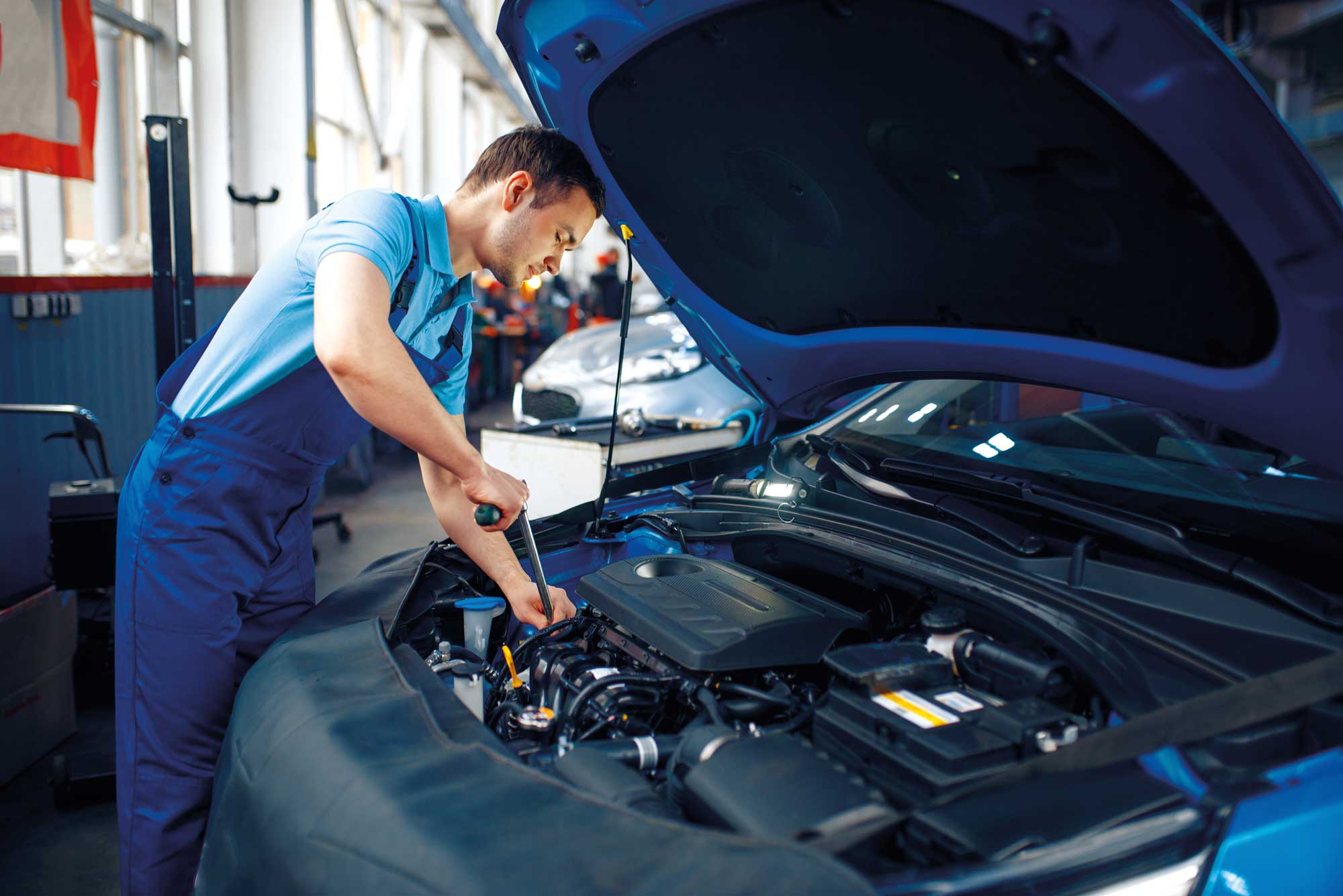 Meccanico al lavoro in officina mentre ripara il motore di un’auto con il cofano aperto, utilizzando un attrezzo manuale.