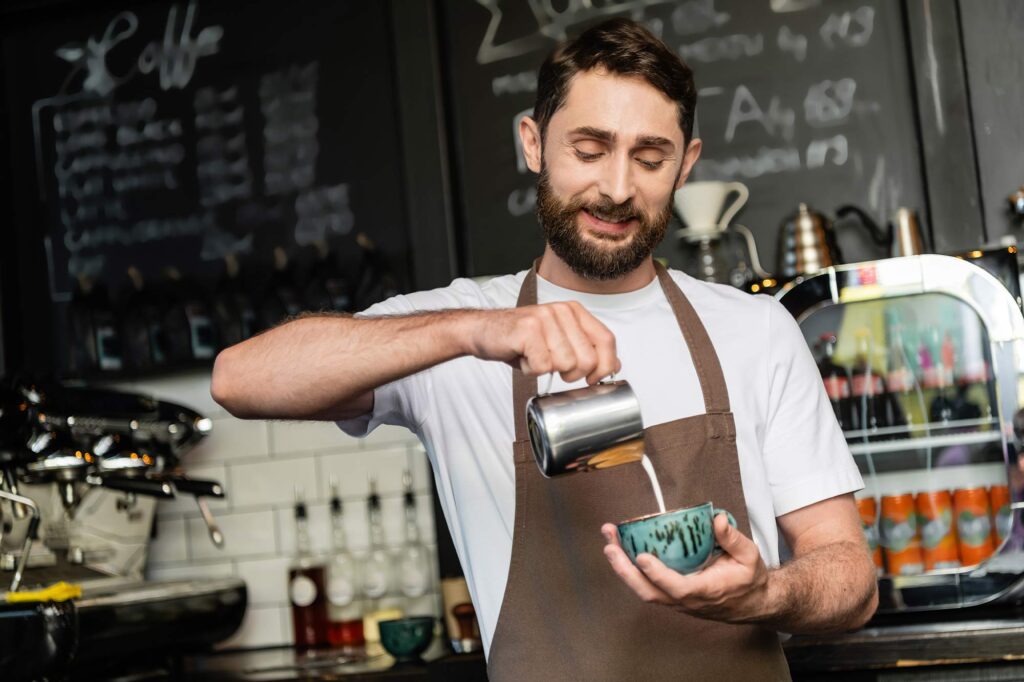 Barista che versa il latte in una tazza per preparare un cappuccino in una caffetteria, sorridendo mentre lavora dietro al bancone.